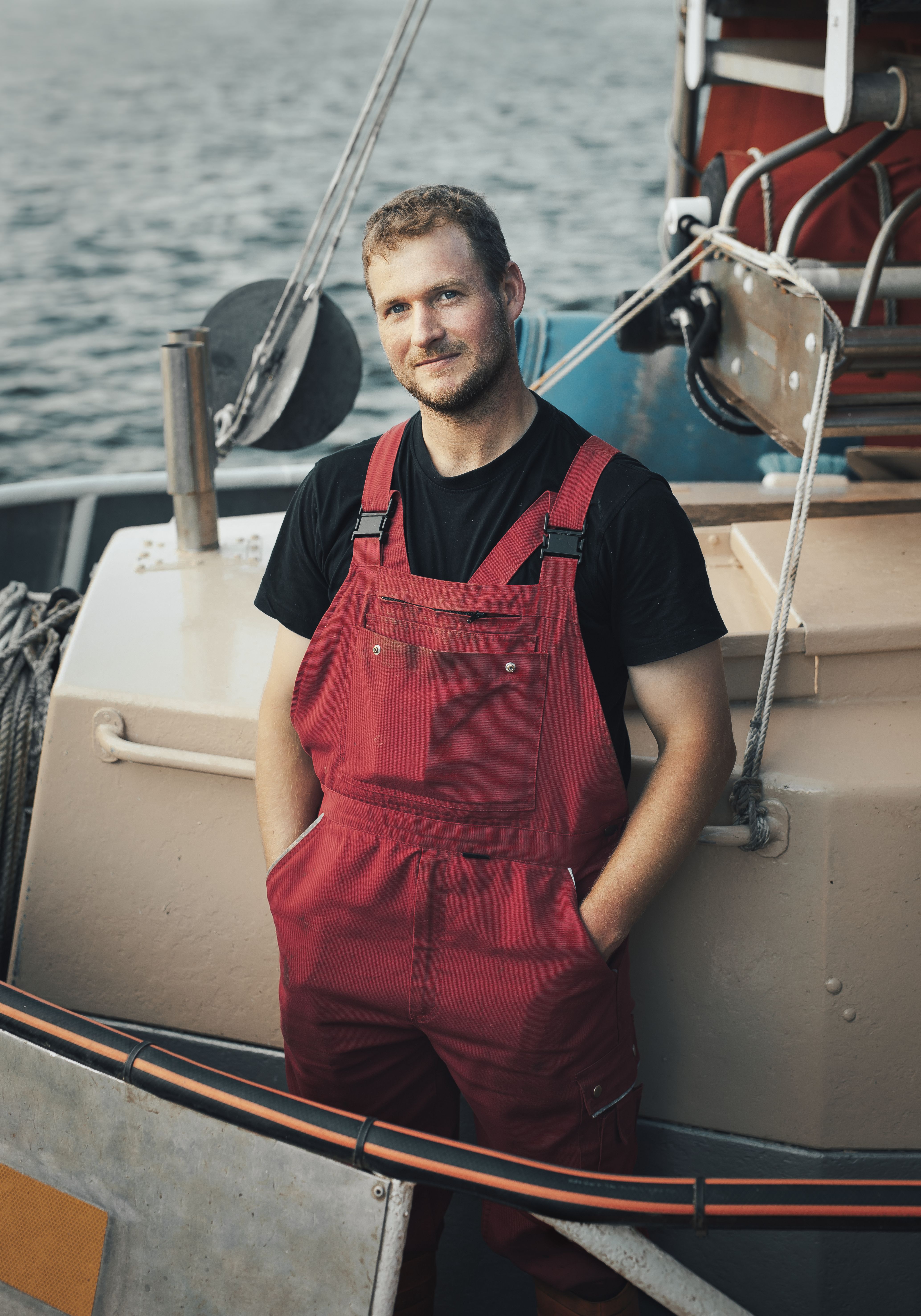 Ein junger Mann steht auf einem Boot. Er trägt eine Latzhose und sieht in die Kamera. 