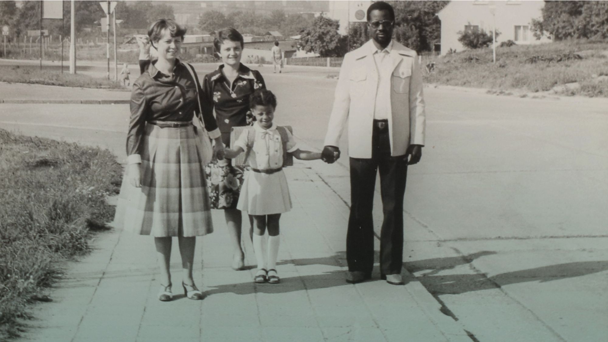 Zwei Frauen links im Bild lächeln in die Kamera. Ein kleines Mädchen rechts von ihnen hält die Hand einer Frau. Rechts im Bild an ihrer Hand steht ihr Vater mit Sonnenbrille. 