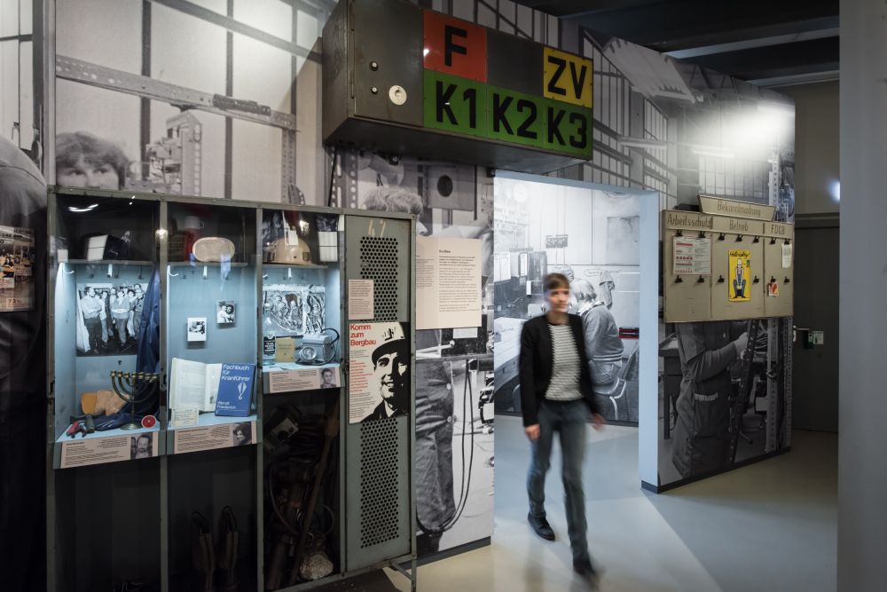 A young woman walks into an exhibition room; on her left four lockers serve as show cases