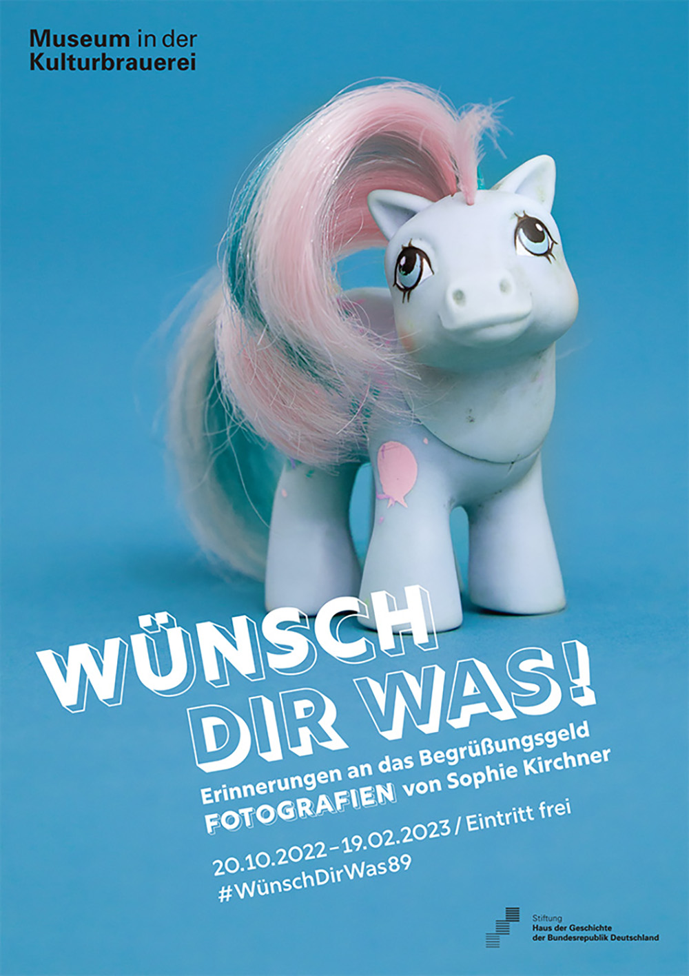 Plakat: Wünsch dir was! Erinnerungen an das Begrüßungsgeld mit Fotografien von Sophie Kirchner. Weißes Spielzeug-Pferdchen mit rosa-hellblauer Mähne auf blauem Hintergrund.
