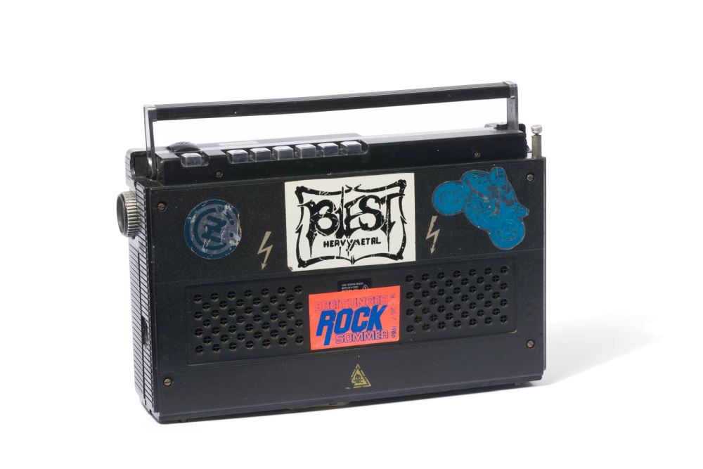 Tragbarer kleiner schwarzer Stern-Radiokassettenrecorder mit diversen Stickern, unter anderem: Biest Heavy Metal, Breitunger Rock Sommer und zwei Hochspannungs-Pfeile.