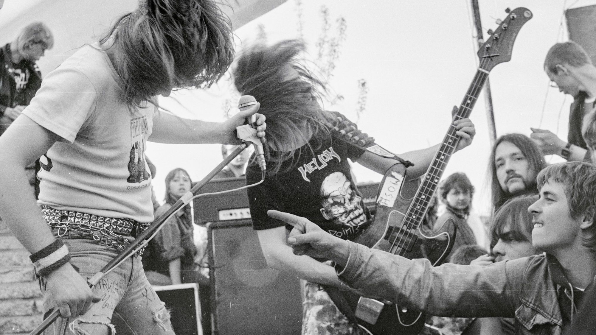 Zwei Personen mit langen haaren stehen erhöht zwischen anderen Menschen, eine hält einen Mikrofonständer, einer eine E-Gitarre. Beide schwingen ihre Haare und sind in Bewegung. Um sie herum sind junge Leute zu sehen, die zum Teil zu den Musikern, zum Teil in die Kamera schauen.