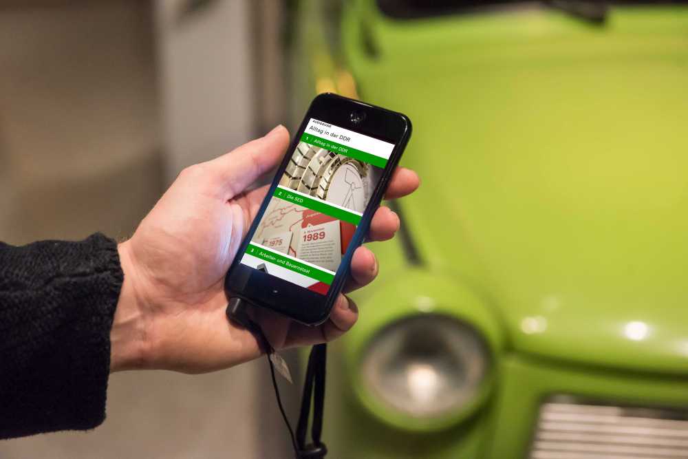 Hand mit Smartphone, auf dem Display zu sehen ist der AudioGuide zur Dauerausstellung 'Alltag in der DDR' im Museum in der Kulturbrauerei Berlin