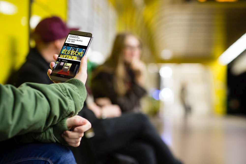 Hand mit Smartphone, auf dem Display zu sehen ist der AudioGuide mit Audiodeskription zur Wechselausstellung 'Deutsche Mythen seit 1945' im Haus der Geschichte Bonn