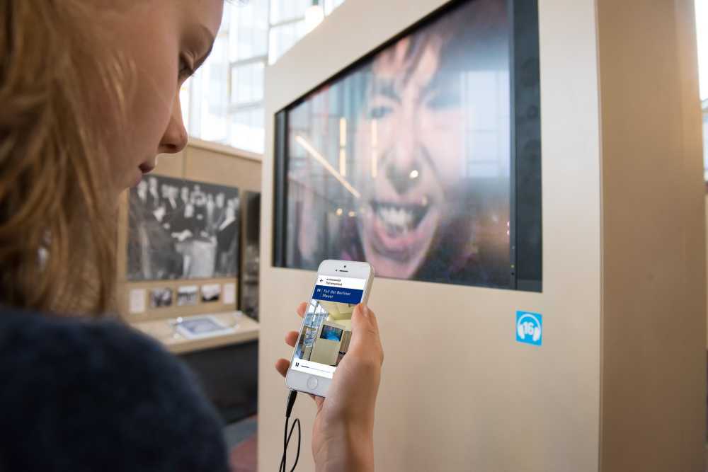 Hand mit Smartphone, auf dem Display zu sehen ist der AudioGuide zur Dauerausstellung 'Ort der deutschen Teilung' im Tränenpalast Berlin