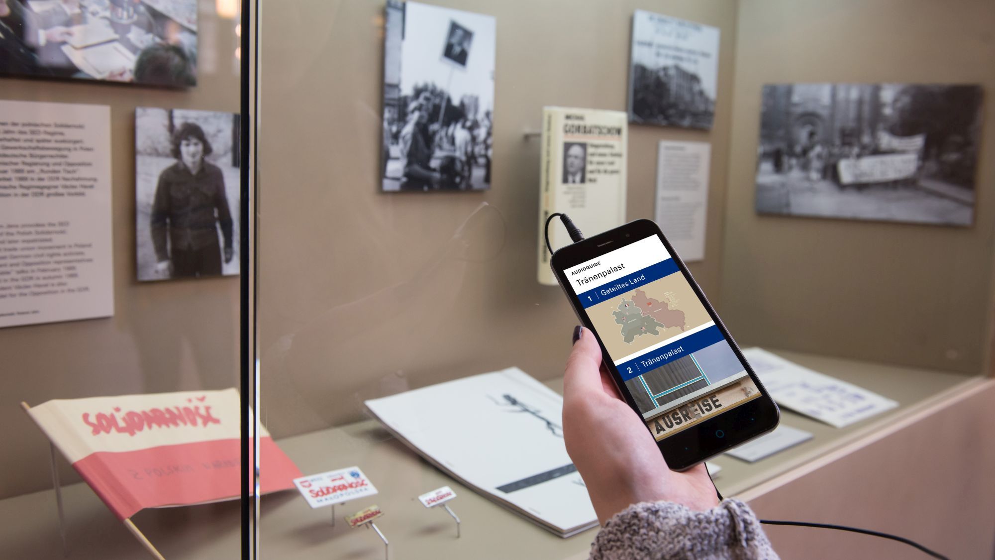 Hand mit Smartphone, auf dem Display zu sehen ist der AudioGuide zur Dauerausstellung 'Ort der deutschen Teilung' im Tränenpalast Berlin