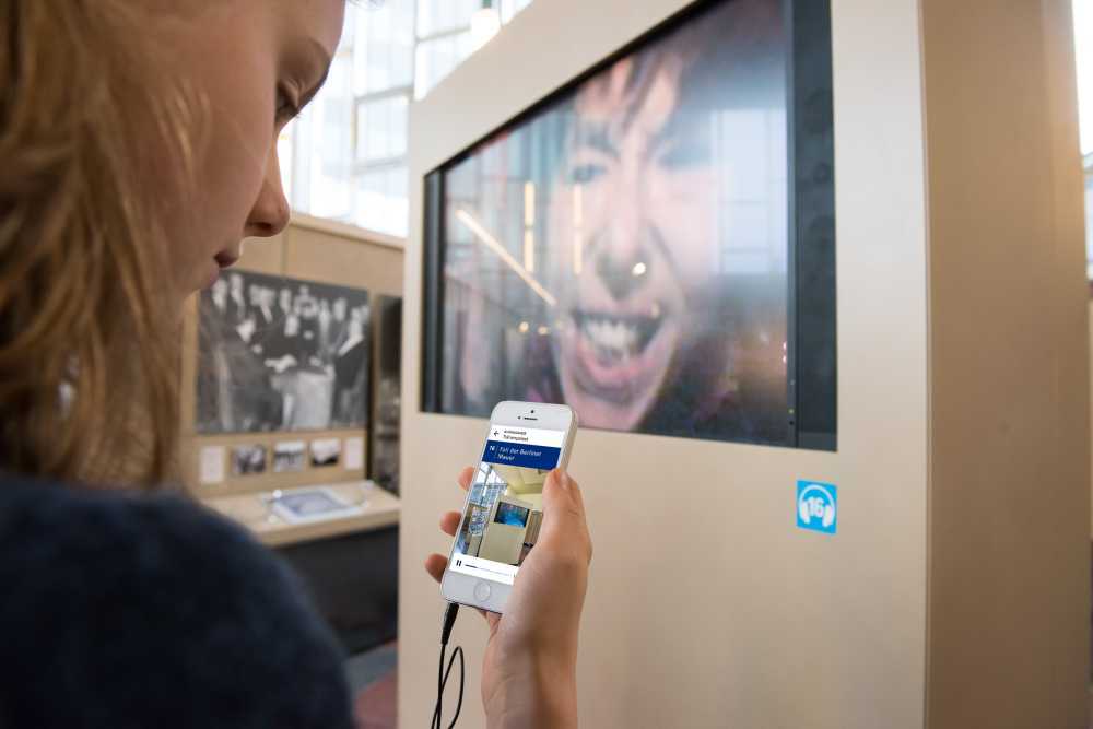 Hand mit Smartphone, auf dem Display zu sehen ist der Audioguide zur Dauerausstellung 'Ort der deutschen Teilung' im Tränenpalast Berlin