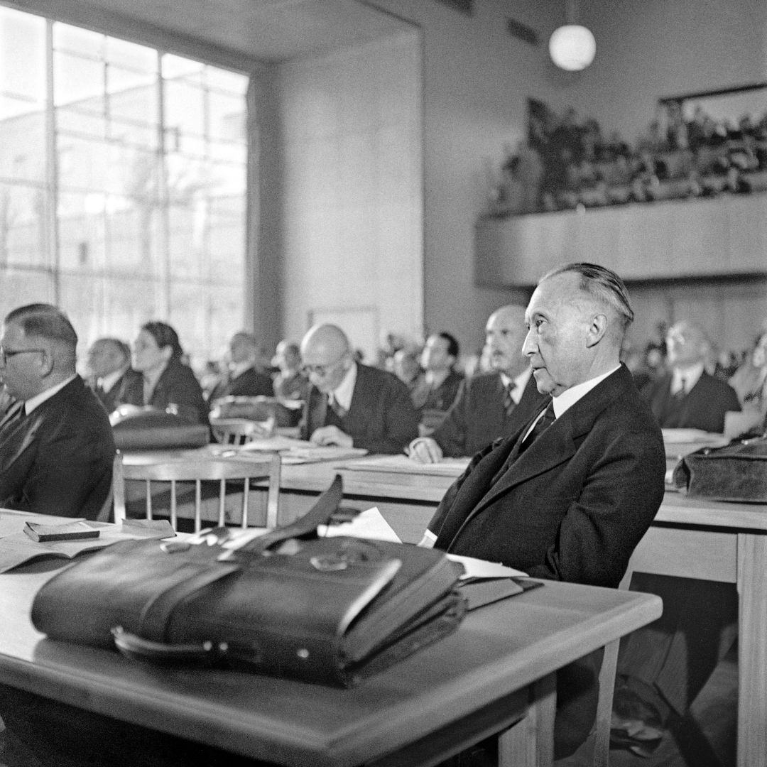 Konrad Adenauer, in älterer Mann im Anzug, sitzt auf diesem historischen Foto an einem Tisch, vor ihm liegt seine Aktentaschen. Hinter und neben ihm sitzen weitere Personen an in Reihen aufgestellten Tischen.