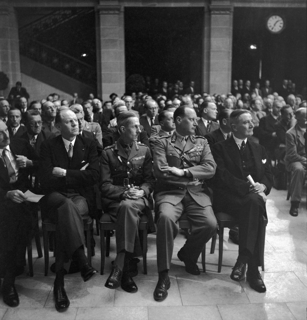 Auf dem historischen Foto sitzen zwei Männer in militärischen Uniformen in der ersten von mehreren Stuhlreihen zwischen vielen anderen Männern in Anzügen in einem großen Saal.