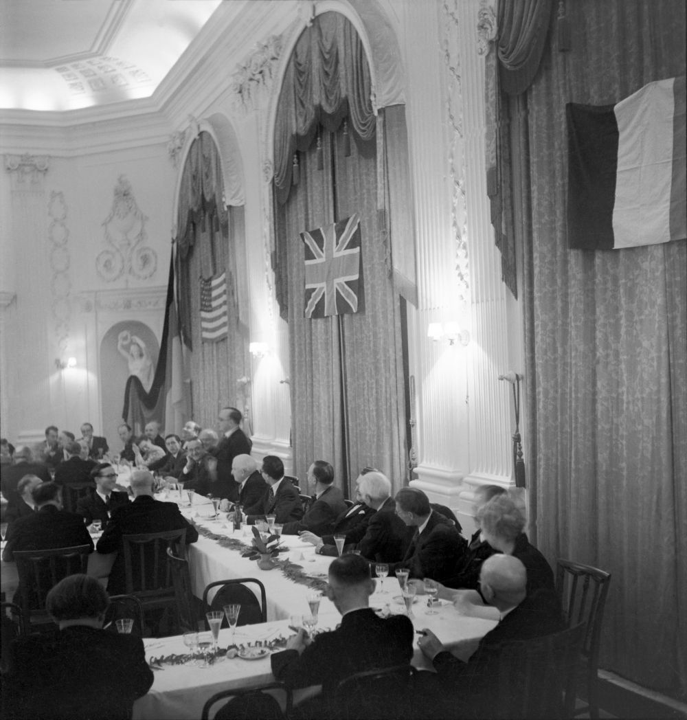 Auf dem historischen Foto sitzen Männer in anzügen an einer Festtafel und schauen zu einem stehenden Redner hin. Die Flaggen der USA, Großbritanniens und Frankreichs sind an Vorhängen in dem hohen Saal angebracht.