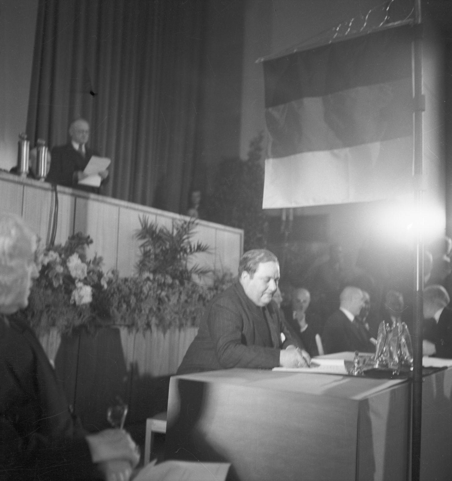 Auf dem historischen Foto sitzt Carlo Schmid im Anzug an einem Tisch und hält einen Stift in der Hand. Über ihm ist eine Deutschlandfahne angebracht. 