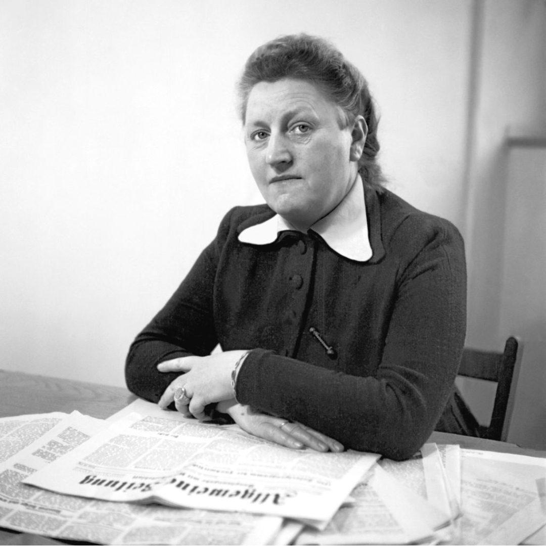 Auf dem historischen Foto sitzt Elisabeth Selbert, eine Frau mittleren Alters an einem Tisch und blickt direkt in die Kamera. Vor ihr liegt eine ausgebreitete Zeitung. Sie trägt ein dunkles Kleid mit weißem Kragen und hat die dunklen Haare zusammengebunden.