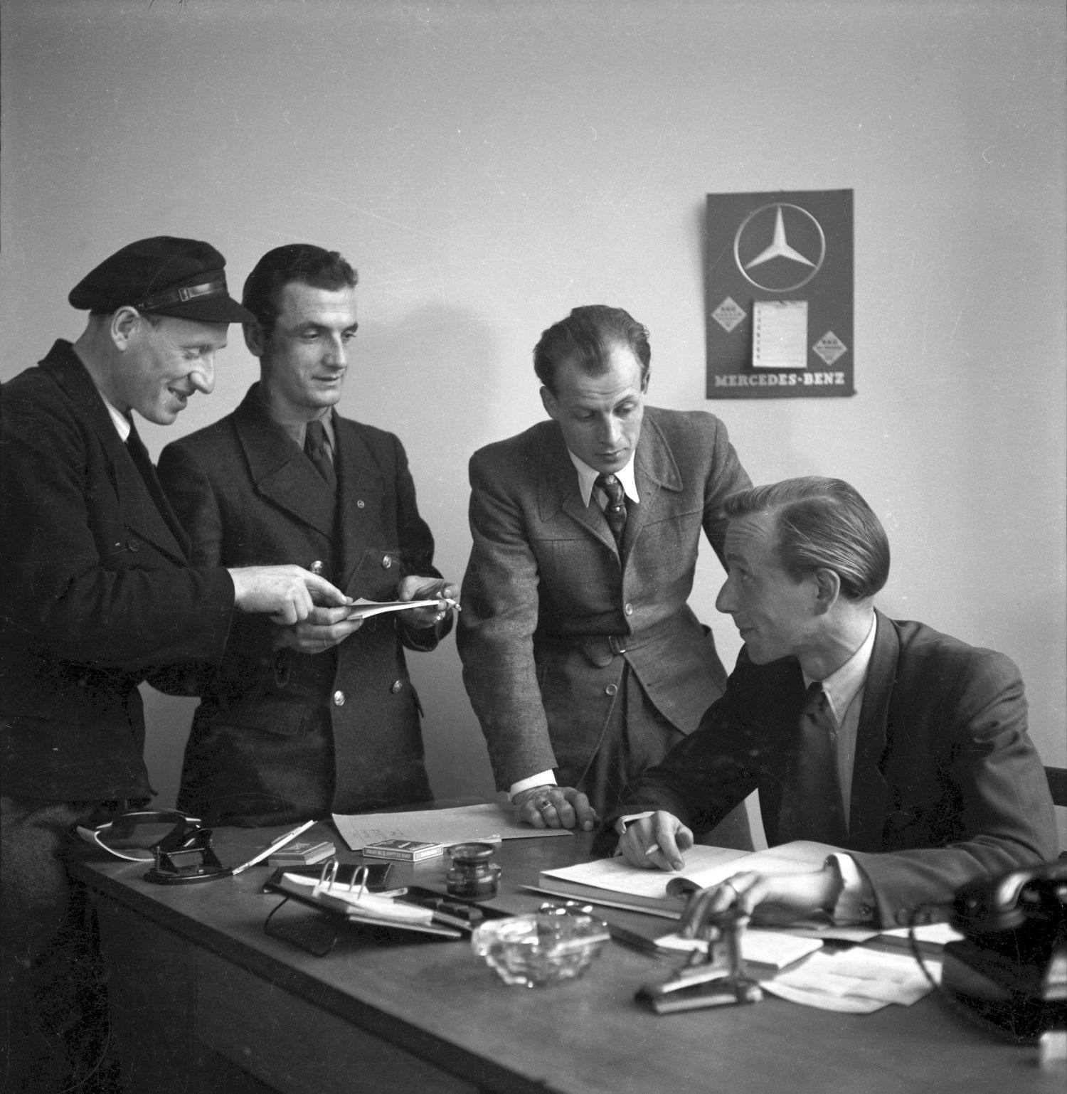 Auf dem historischen Foto sitzt ein Mann im Anzug an einem Schreibtisch, drei weitere stehen neben ihm. Sie scheinen etwas zu besprechen.