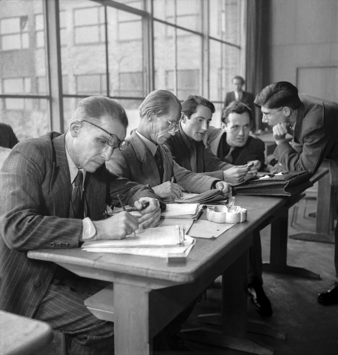 Auf dem historischen Foto sitzen vier Männer in Anzügen an einem Tisch. Einer schreibt, einer liest und zwei unterhalten sich mit einem weiteren Mann, der neben dem Tisch steht und sich zu ihnen herunterbeugt.