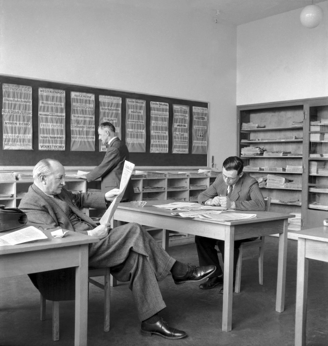 Auf dem historischen Foto lesen zwei Männer in Anzügen an einem Tisch sitzend Zeitung. Im Hintergrund steht ein weiterer Mann an einem Regal mit vielen offenen Fächern und Papierstapeln oder Zeitungen.