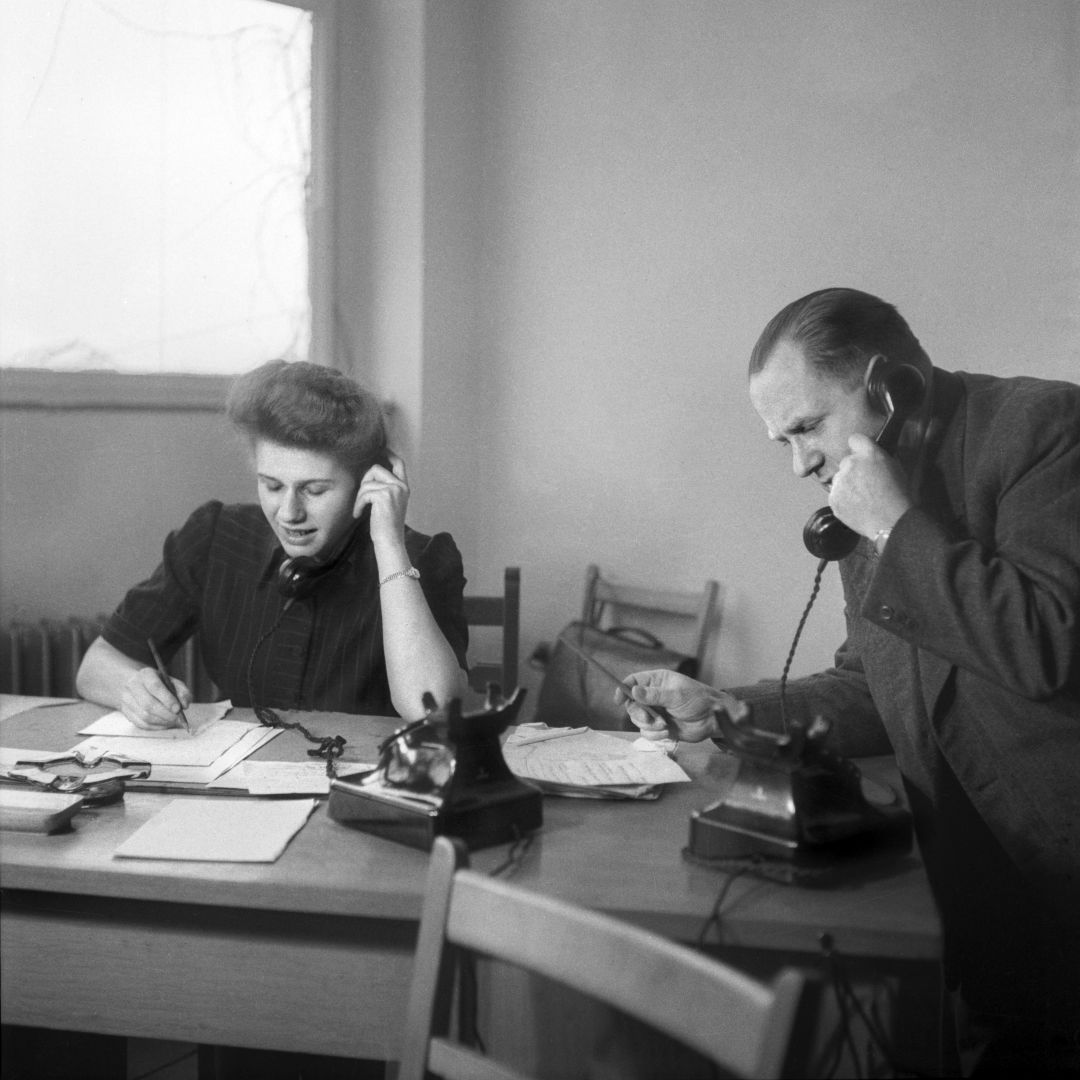 Auf dem historischen Foto haben eine Frau und ein Mann Telefonhörer am Ohr. Sie sitzt an einem Tisch und macht sich Notizen, er steht nebem dem Tisch und hält einen Stift in seiner rechten Hand.