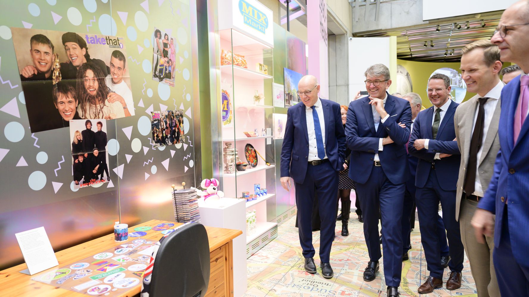 Wolfram Weimer schaut sich mit einer kleinen Gruppe Männer einen Ausstellungsbereich zu den 1990er-/frühen 00er-Jahren an. An der Wand hängen Poster, u.a. von der Boyband "Take That".