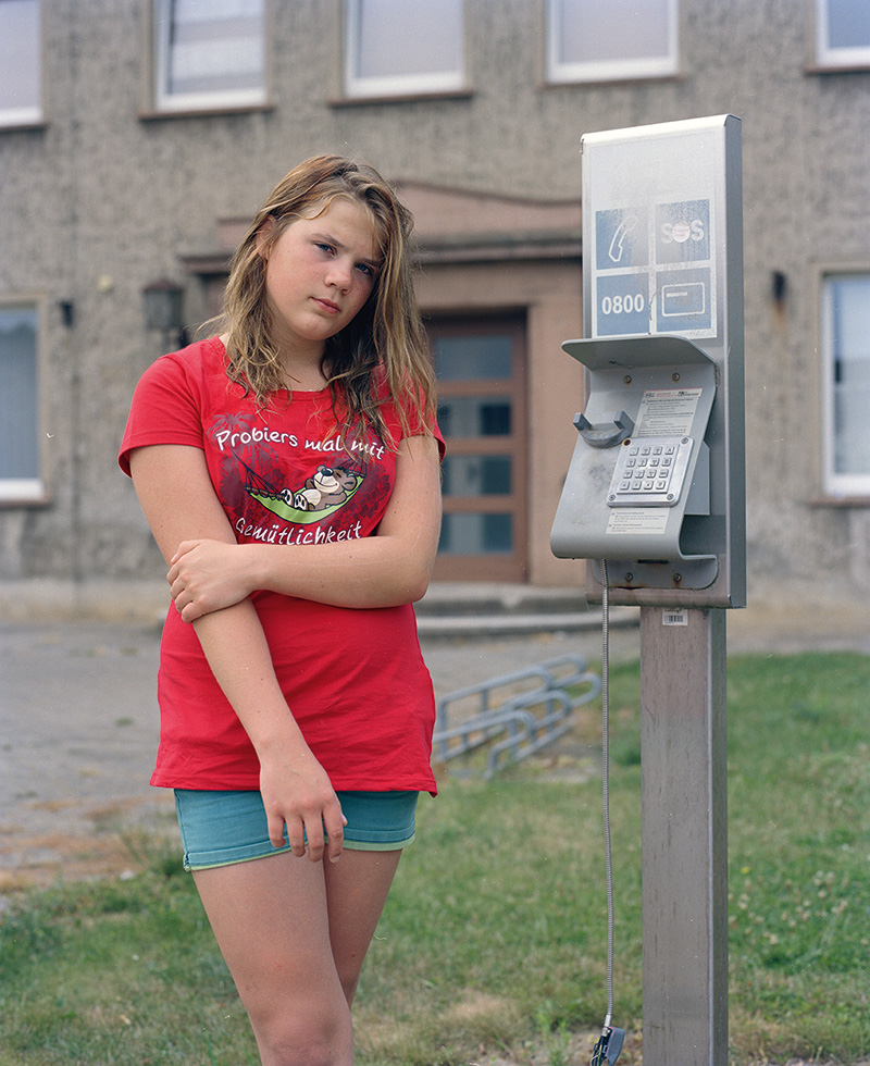 Ein Mädchen steht vor einem grauen Haus, neben ihr befindet sich eine sich offensichtlich nicht in Betrieb befindende Telefonstation.