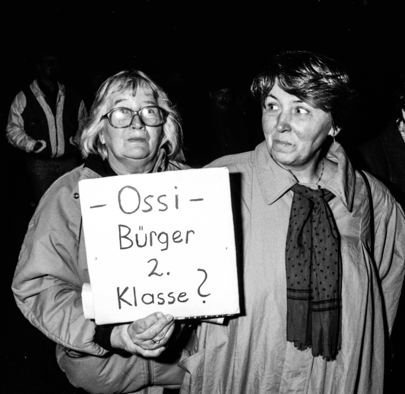 Zwei Frauen, eine trägt ein Schild mit der Aufschrift -Ossi- Bürger 2. Klasse?