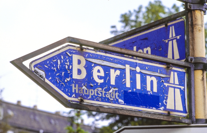 Ein Straßenschild mit Hinweis auf die Autobahn nach Berlin, unter dem Wort Berlin steht Hauptstadt, dahinter ist etwas überklebt.