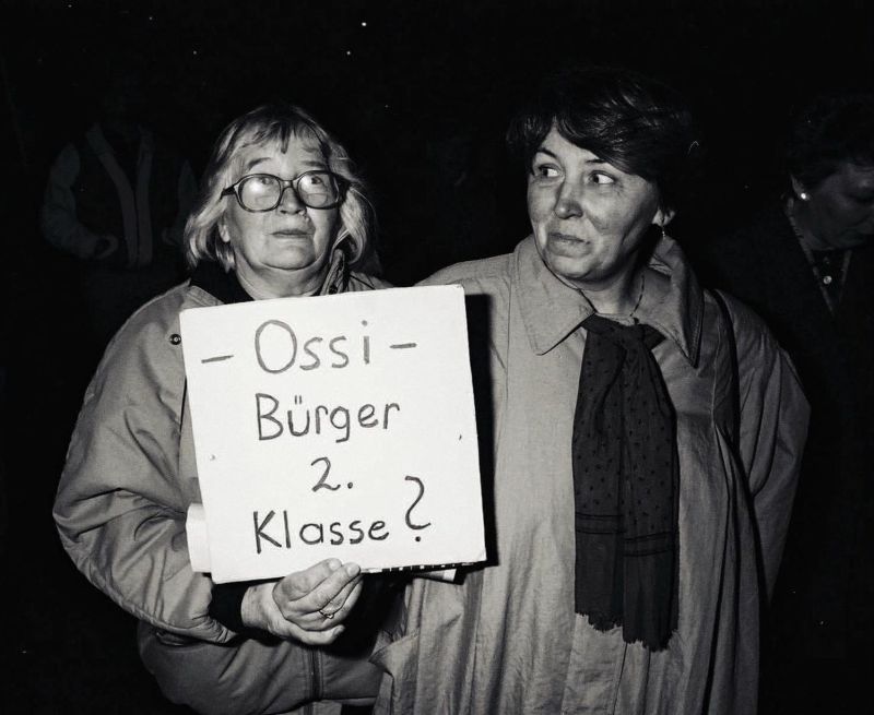 Zwei Frauen, eine davon trägt ein Plakat mit der Aufschrift - Ossi - Bürger 2. Klasse?