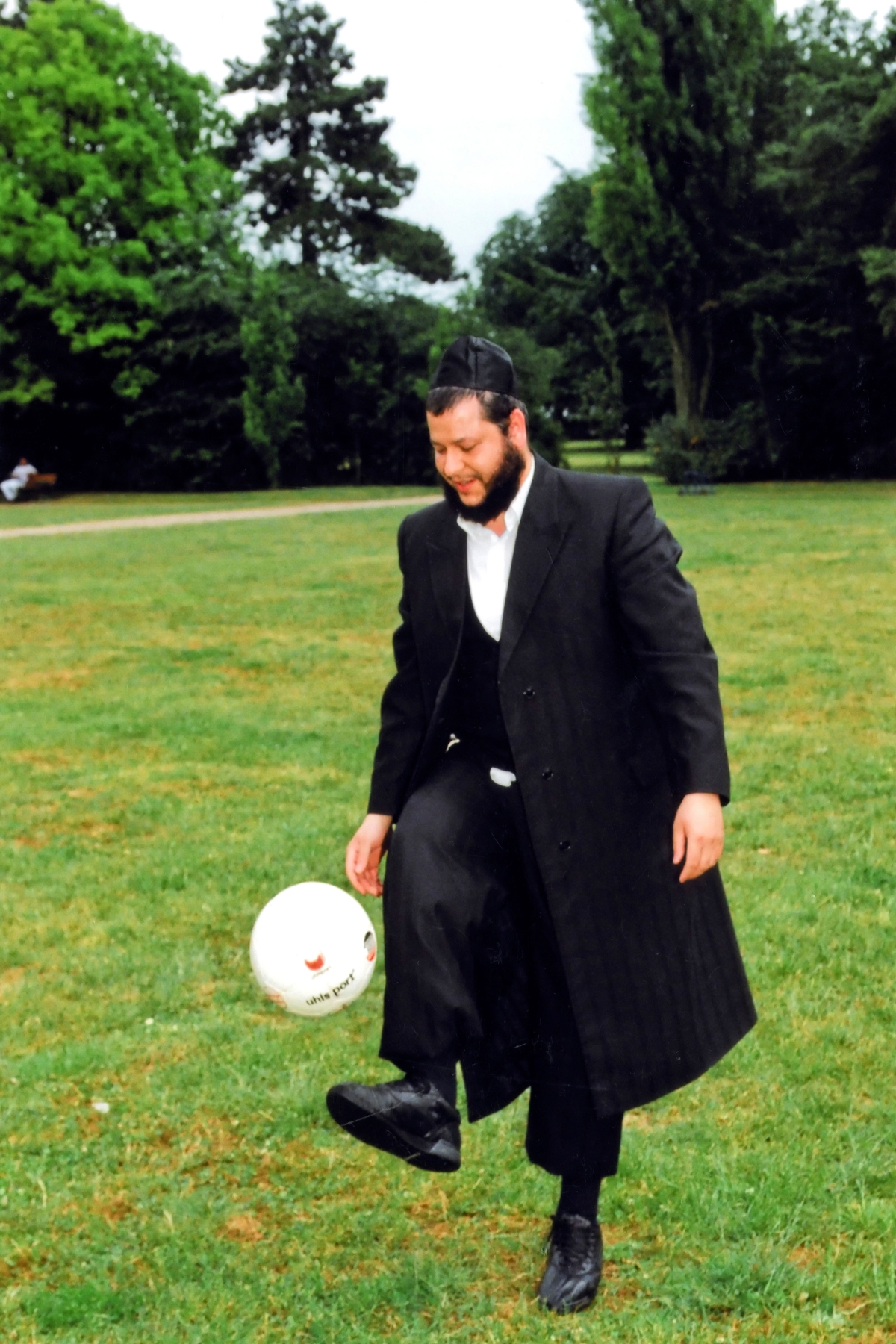 Ein Mann mit schwarzem Anzug, Mantel und Kippa spielt auf einer Wiese mit einem Fußball.