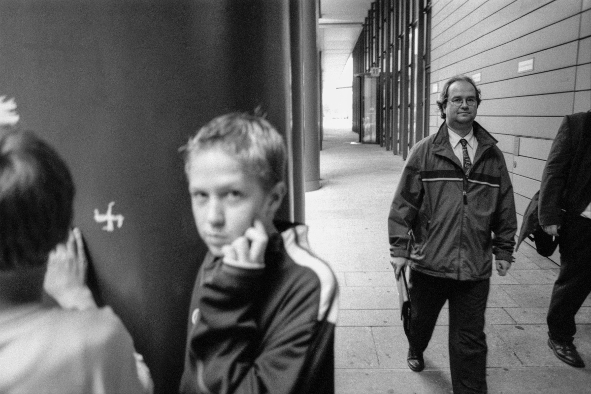 Ein Junge blickt den Betrachter direkt an, ein weiterer ritzt gerade etwas in Beton, zwischen ihnen ein Hakenkreuz, aus dem Hintegrund kommen zwei Erwachsene.