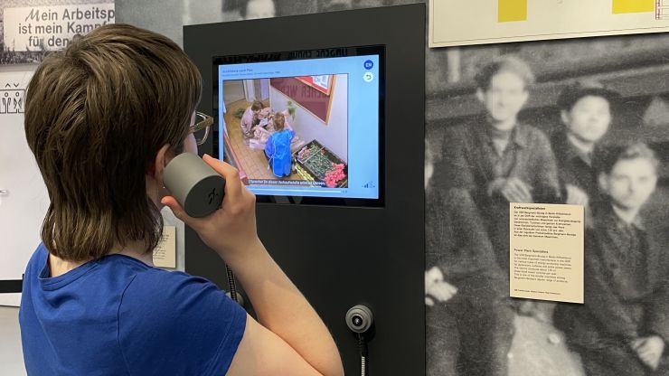Frau steht in der Ausstellung mit Hörer einer Medienstation am Ohr