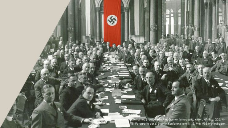 Historisches Foto: Männer sitzen an langen Tischen, im Hintergrund hängt ein Banner mit Hakenkreuzsymbol 
