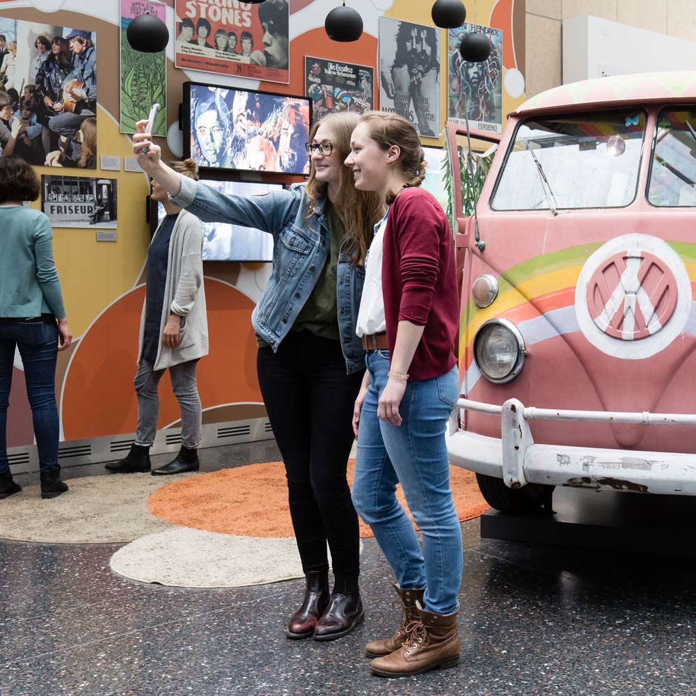 Zwei junge machen ein Selfie vor einem rosafarbenen VW-T1-Bus, auf dessen Front ein Regenbogen und ein Peace-Zeichen aufgemalt sind.