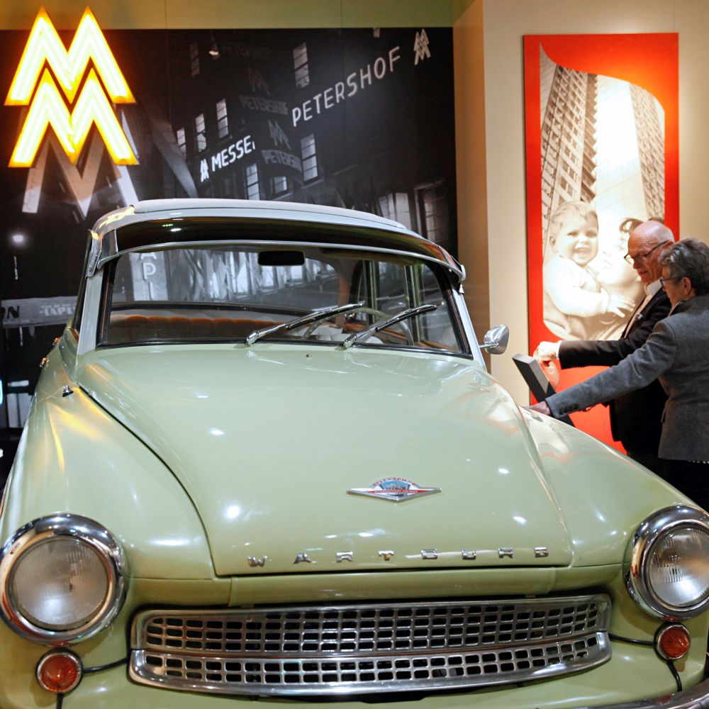 In einem Ausstellungsraum steht ein frontal fotografiertes, hellgrünes Auto mit chromfarbenen Scheinwerferblenden und Kühlergrill. Daneben stehen zwei Besucher.