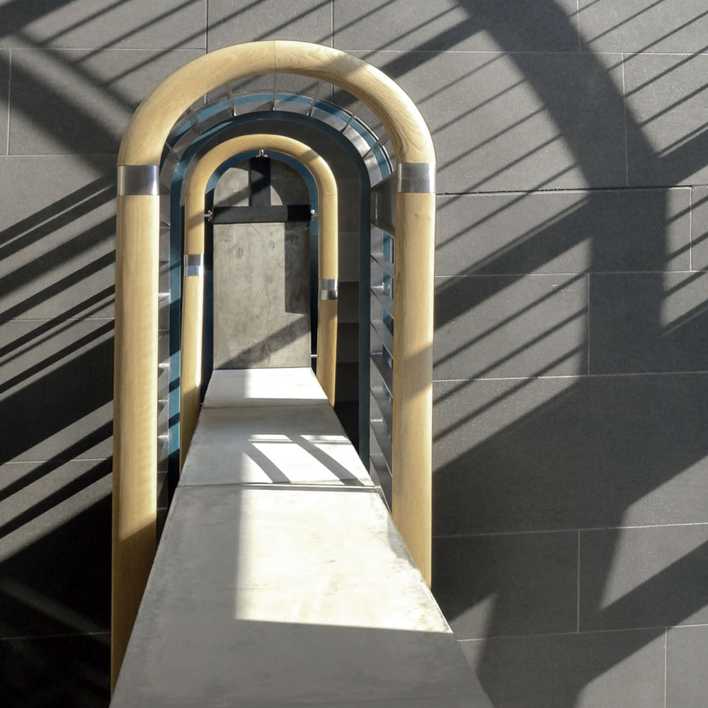 Eine schmale Betonbrücke läuft auf eine Tür aus Sichtbeton zu. Über Brücke und Tür spannen sich helle Holzbögen. An der Wand zeichnen sich geometrische Schatten ab.
