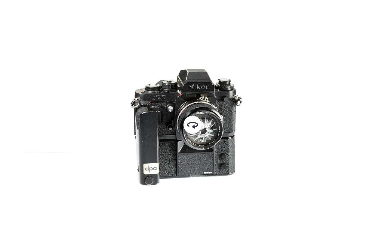 Schwarze Nikon F3T Spiegelreflexkamera. Unten Motorgehäuse, Handgriff rechts. Kamera für professionellen Einsatz. Aufschrift Vorderseite: Nikon F3T, dpa (Aufkleber), Rückeseite: Nikon Motor Drive Wattenberg (Aufkleber). Stark beschädigtes Objektiv und zersplitterte Linse.