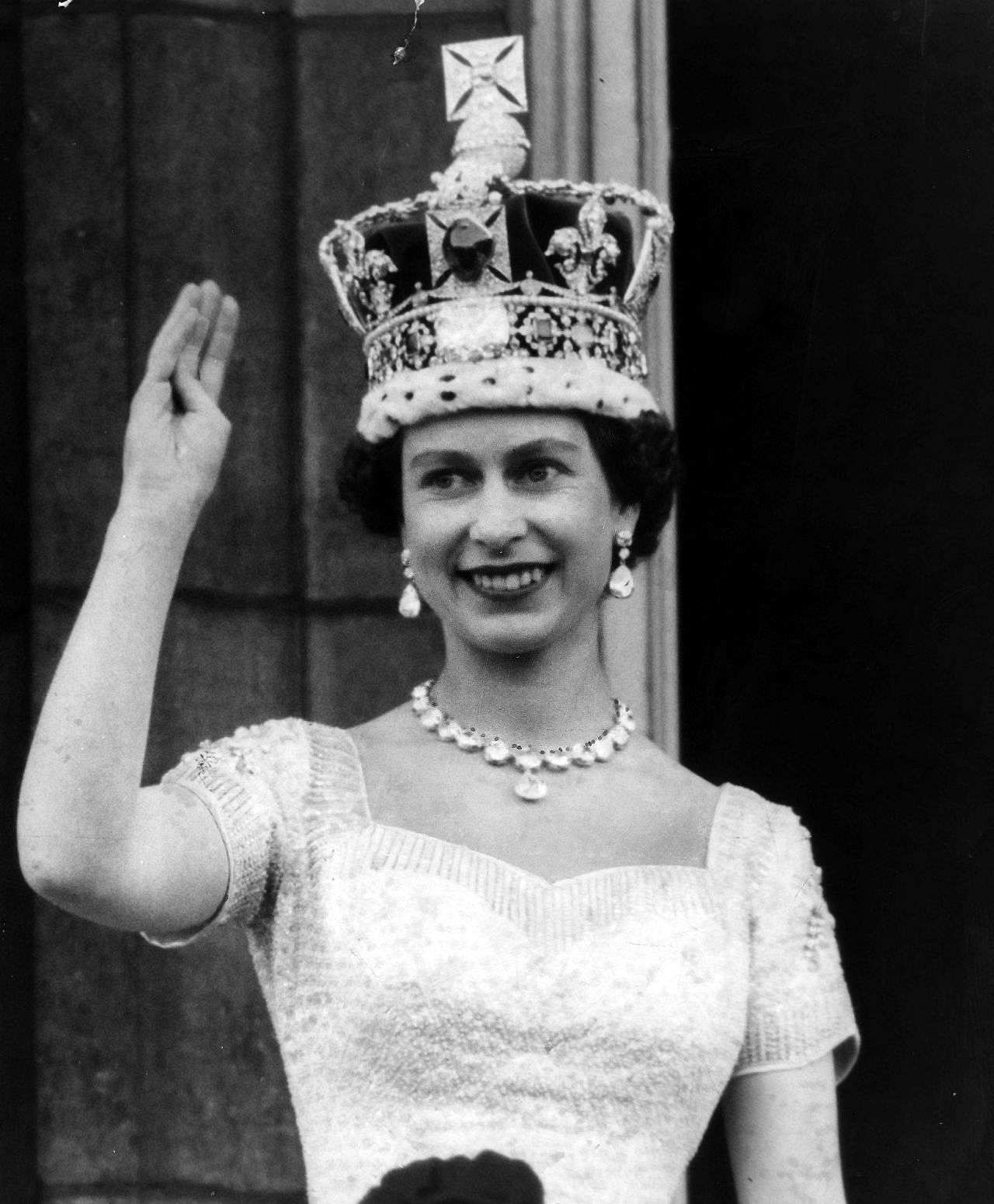 Elisabeth II winkt mit erhobenen Arm, sie trägt ein helles Kleid und eine Krone.
