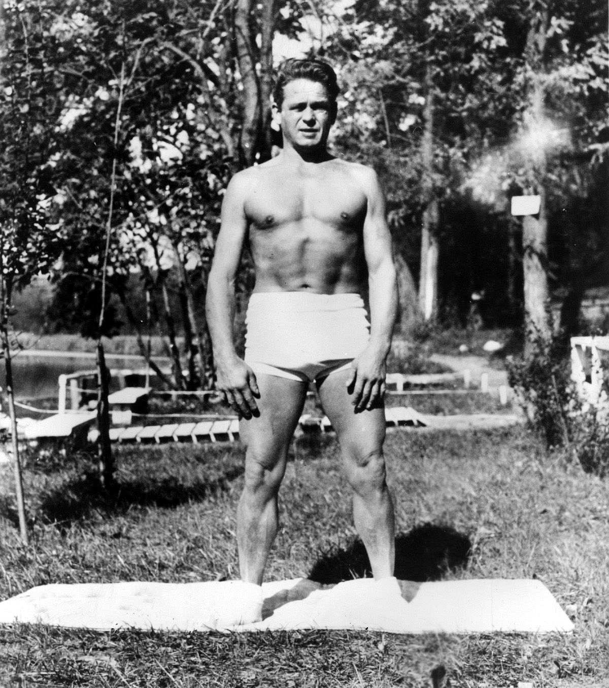 Schwarz-weiße Fotografie von Joseph Pilates. Er steht draußen im Garten breitbeinig auf einer Turnmappe und trägt nur eine weiße Shorts. Sein Körper ist muskulös und er schaut mit zusammengekneiften Augen in die Kamera.