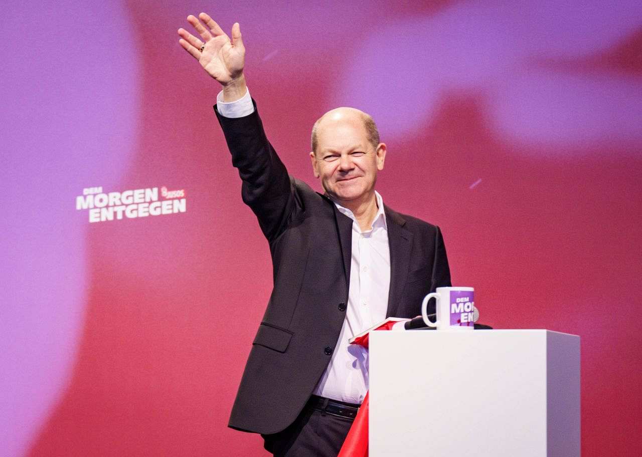 Olaf Scholz steht vor einer roten Wand und winkt lächelnd. Er trägt dabei einen schwarzen Anzug, vor ihm steht eine Tasse auf dem weißen Rednerpult. 