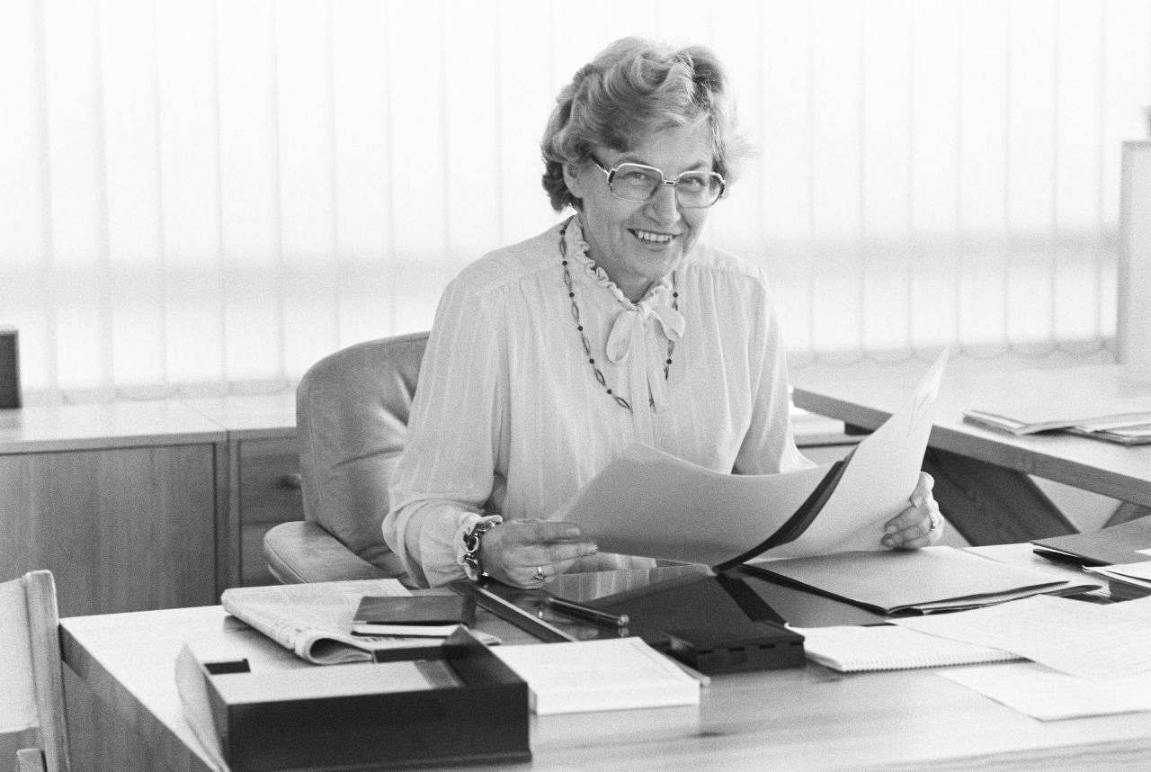 Schwarz-weiße Fotografie von Dorothee Wilms hinter ihrem Schreibtisch. Sie hält eine Aktenmappe in der Hand und lächelt in die Kamera.