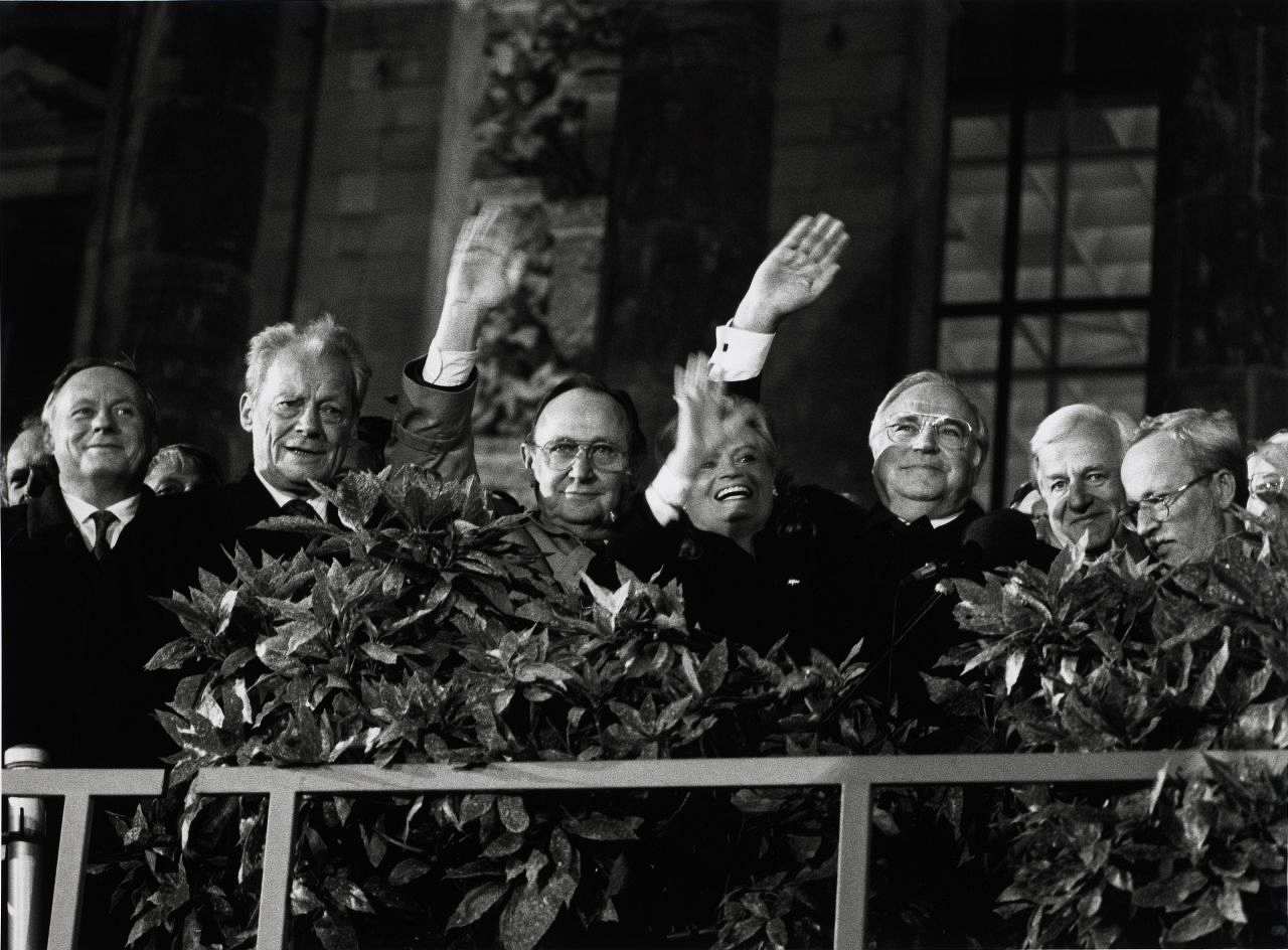 Schwarzweiß-Foto, auf einer Tribüne vor dem Reichstagsgebäude: Oskar Lafontaine, Willy Brandt, Hans-Dietrich Genscher winkend, Hannelore und Helmut Kohl winkend, Richard von Weizsäcker, Lothar de Maizière. Zwischen Geländer und Personen teils Büsche.