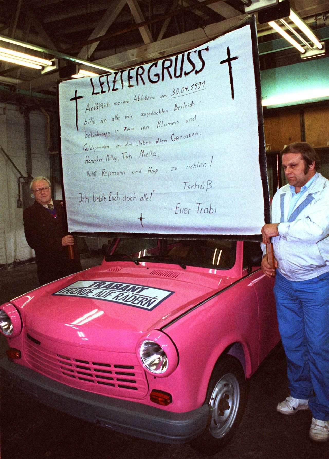 Farb-Foto,  zwei Männer halten ein Transparent in Höhe der Windschutzscheibe über den letzten Trabi, Schriftzug: ‚Letzter Gruss. Anläßlich meines Ablebens am 30.04.1991…‘.  Trabi ist pinkfarben, auf der Motorhaube Aufkleber ‚Trabant – Legende auf Rädern‘.