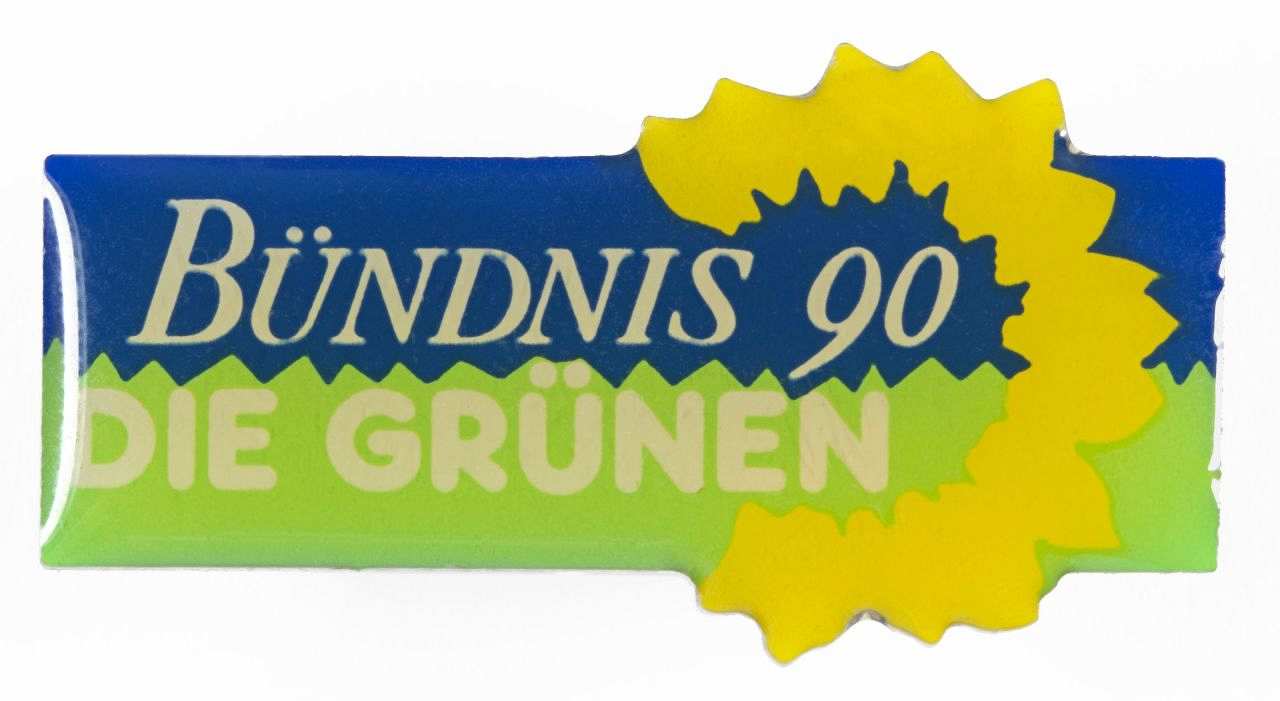 Rechteckiger Anstecker, die obere Hälfte ist blau, die untere grün. Darauf steht in weißer Schrift: Bündnis 90 Die Grünen. Um einen Teil der Schrift geht in einem Halbkreis eine gelbe Sonnenblumenblüte.