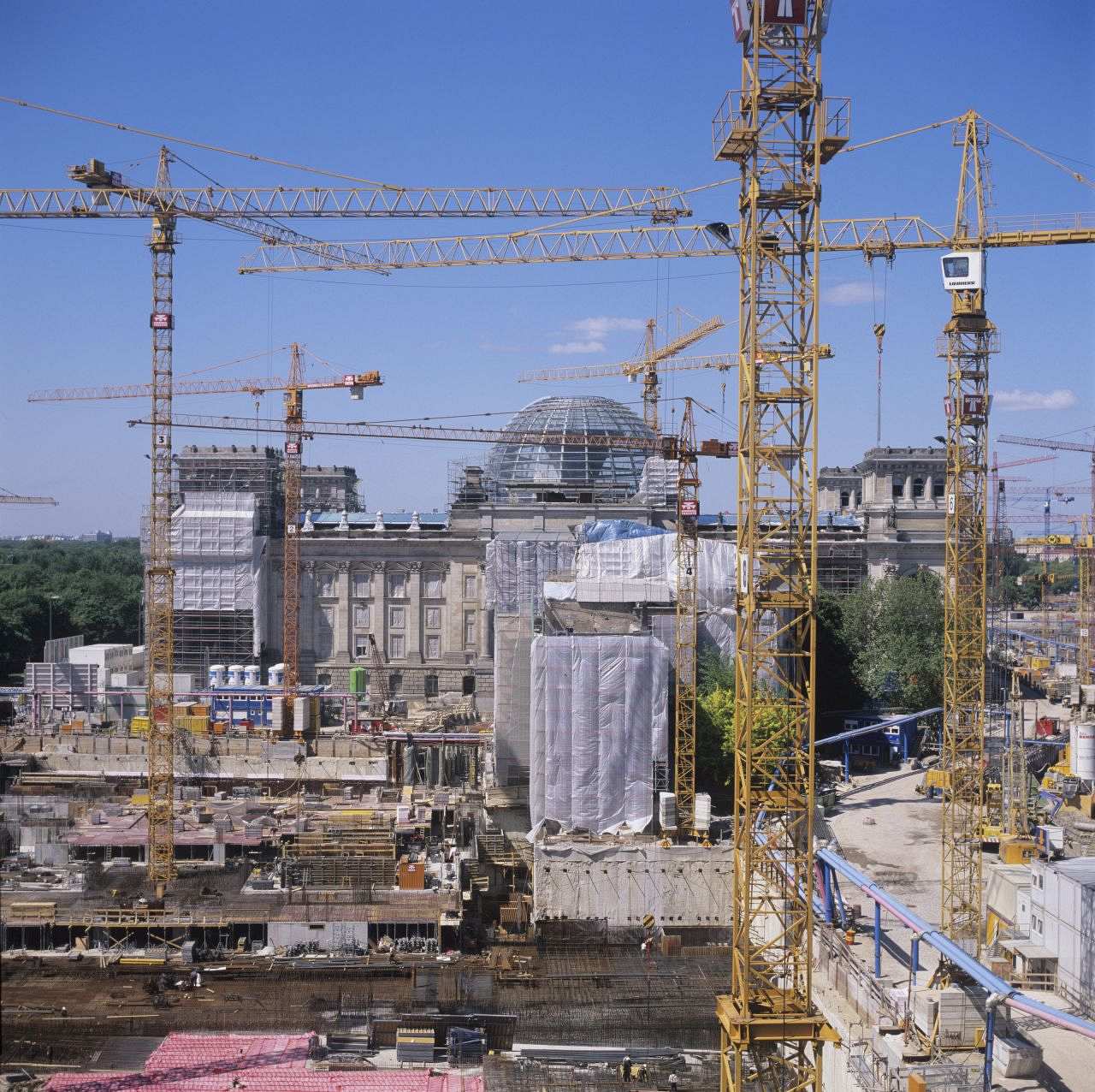 Fotografie einer Baustelle: Im Vordergrund zahlreiche hohe Baukräne, im Hintergrund ist der Reichstag in Berlin zu sehen, der teils mit Baugerüsten und Planen abgedeckt ist.