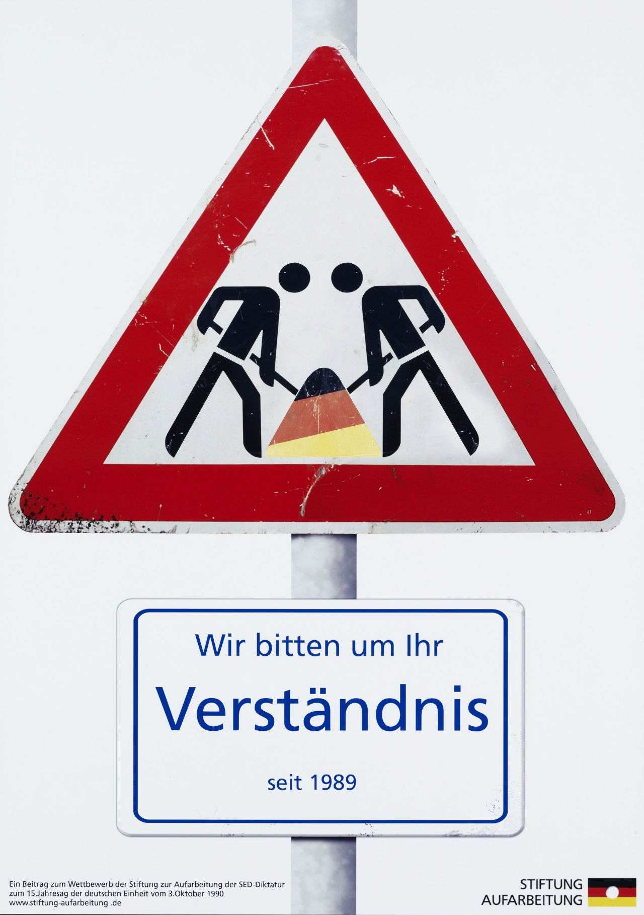 Farbiges Plakat; weißer Hintergrund; dreieckiges Baustellenschild: zwei Strichmännchen mir Schaufeln arbeiten gemeinsam an einem schwarz-rot-goldenen Sandhaufen; unterhalb des Schildes ist weißes Schild mit blauer Schrift angebracht: Wir bitten um Ihr / Verständnis / seit 1989; unten, links, in schwarzer Schrift:  Ein Beitrag zum Wettbewerb der Stiftung zur Aufarbeitung der SED-Diktatur / zum 15. Jahrestag der deutschen Einheit vom 3. Oktober 1990 / www.stiftung-aufarbeitung.de; daneben, rechts: Stiftung / Aufarbeitung, mit Logo.