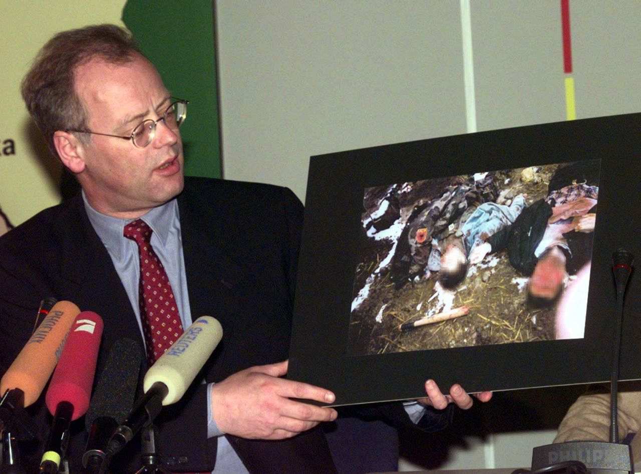 Bundesverteidigungsminister Rudolf Scharping steht links im Bild, vor ihm sind Mikrofone von Journalisten zu sehen. In seiner Hand ein ca. DIN A 3 großes Foto zweier getöteter Kosovo-Albaner.
