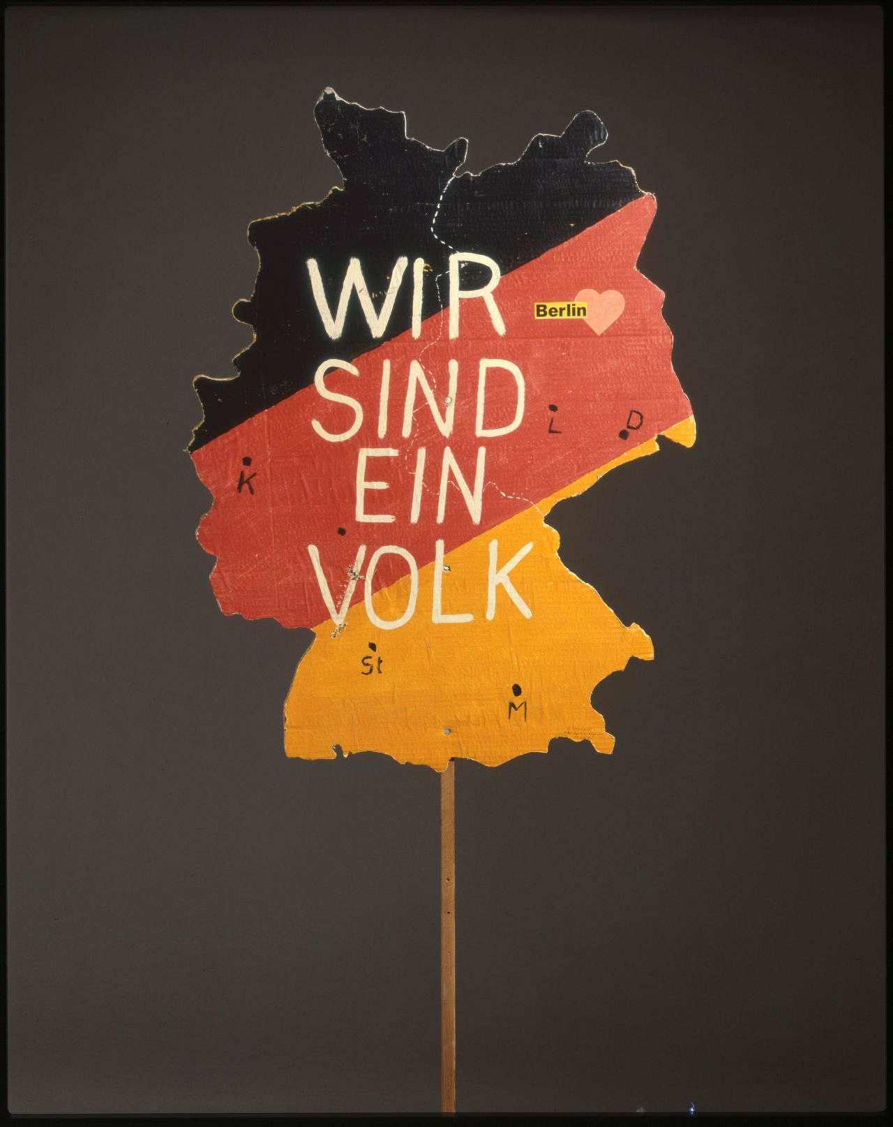 Transparent vom Dezember 1989: Wir sind ein Volk, Transparent in Form des gesamten Deutschlands in den Farben schwarz, rot, gold, darauf in weißer Farbe: Wir sind ein Volk, eingezeichnet sind Dresden, Leipzig, München, Stuttgart, Frankfurt und Köln und jeweils mit den Anfangsbuchstaben gekennzeichnet (außer Frankfurt), Berlin ist in Herzform eingezeichnet und mit vollständigem Namen gekennzeichnet.