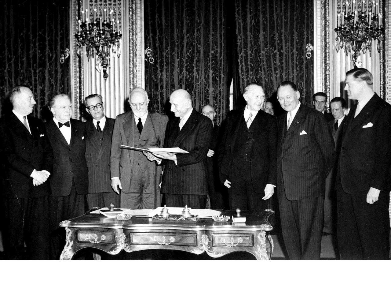 Schwarz-Weiß-Foto. Unterzeichner des EGKS-Vertrags stehen hinter einem reich-verzierten verschnörkelten Tisch. Robert Schuman hält vermutlich den Vertrag in den Händen. Carlo Sforza - neben ihm stehend - schaut hinein.