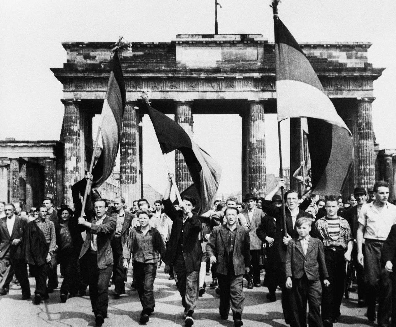 Arbeiter ziehen durch das Brandenburger Tor. Drei von Ihnen tragen Fahnen, von denen zwei als schwarz-rot-golden zu erkennen sind.
