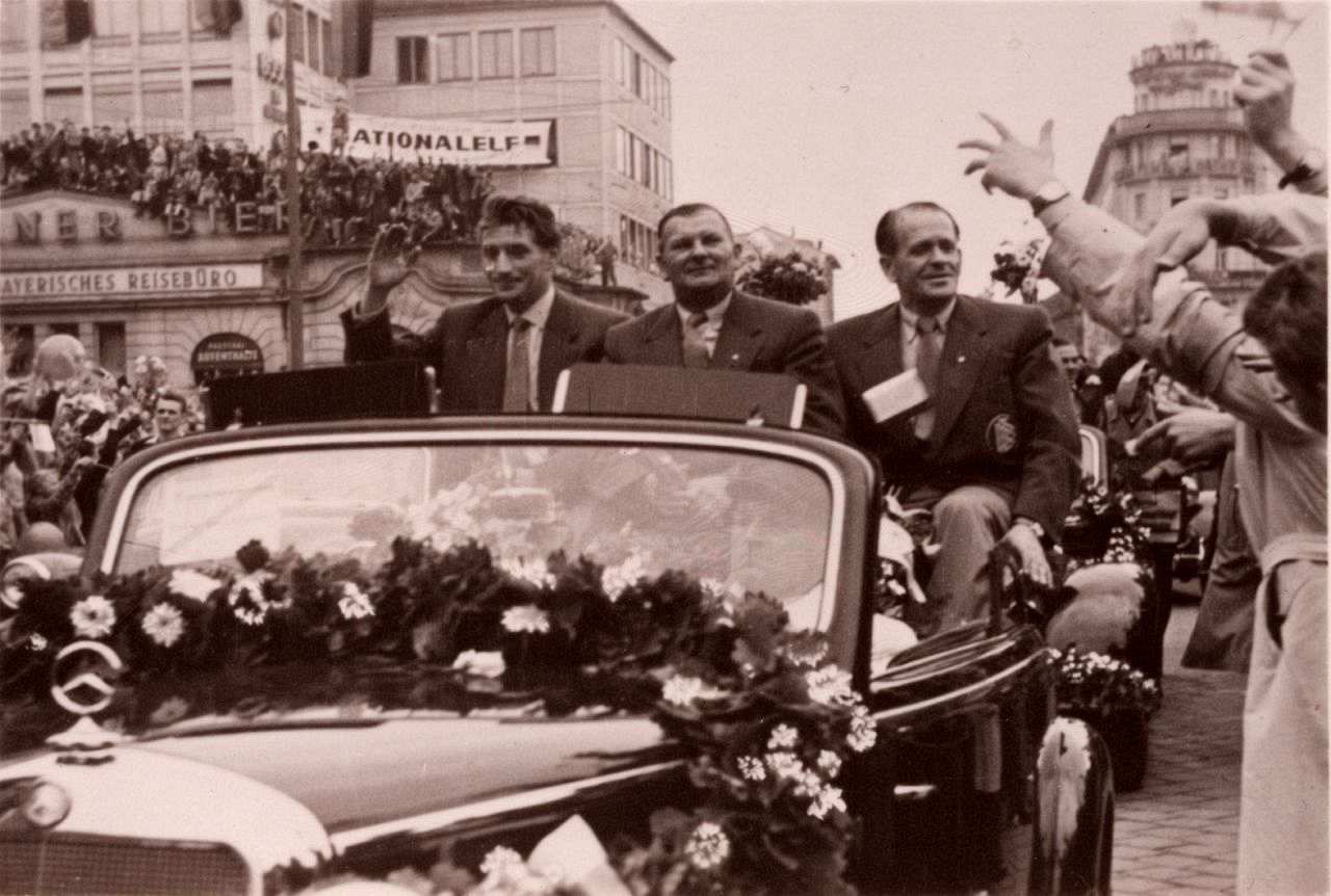Fritz Walter, Hans Huber und Sepp Herberger werden in einem blumengeschmückten Mercedes-Cabriolet, hoch sitzend, durch München gefahren. Zuschauer jubeln ihnen zu.
