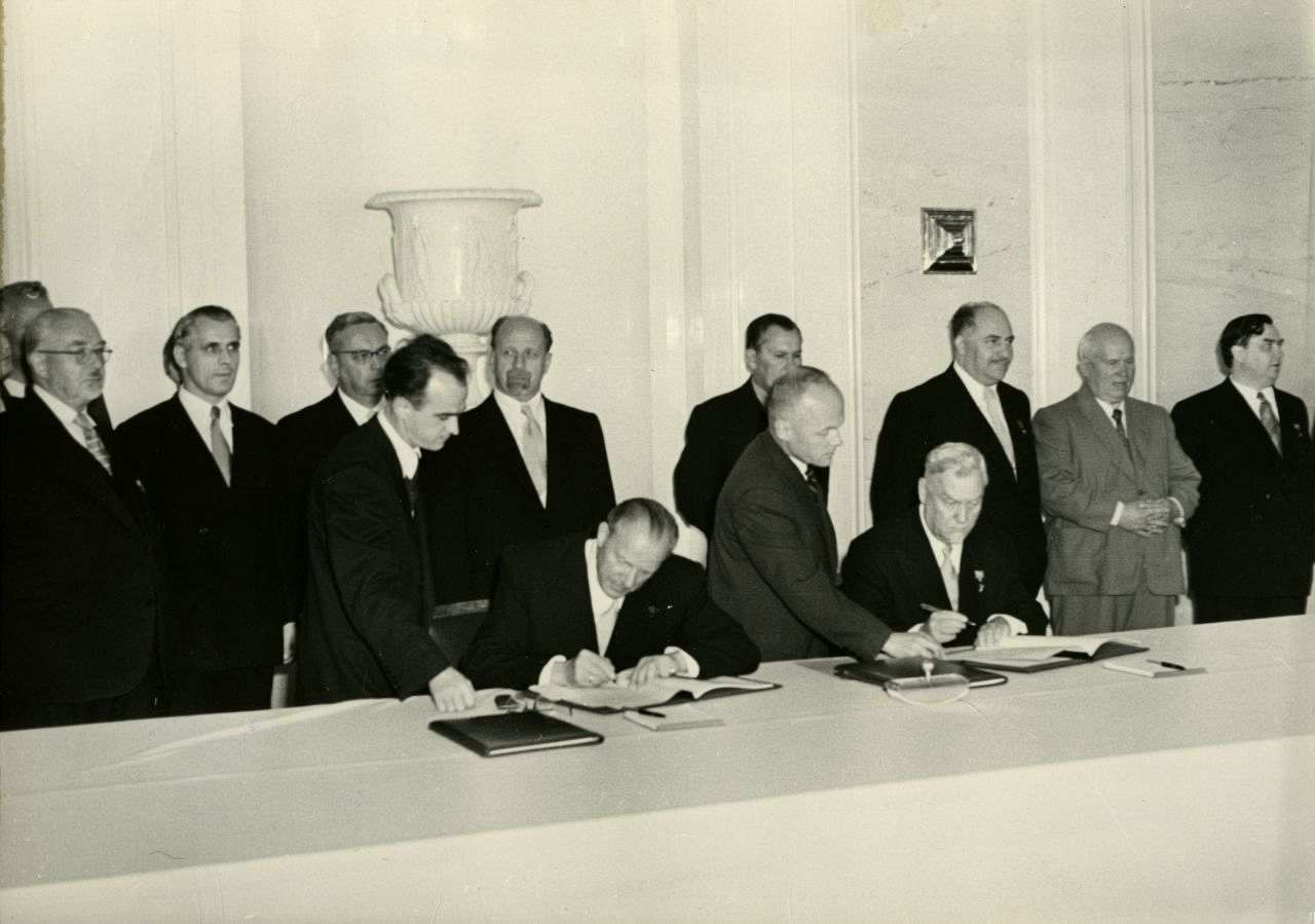 Otto Grotewohl und Nikolai A. Bulganin an einem langen Tisch sitzend unterschreiben den ‚Vertrag über die Beziehungen zwischen der DDR und der UdSSR‘. Hinter ihnen stehend, mehrere weitere der Vertragsunterzeichnung Teilhabende. 