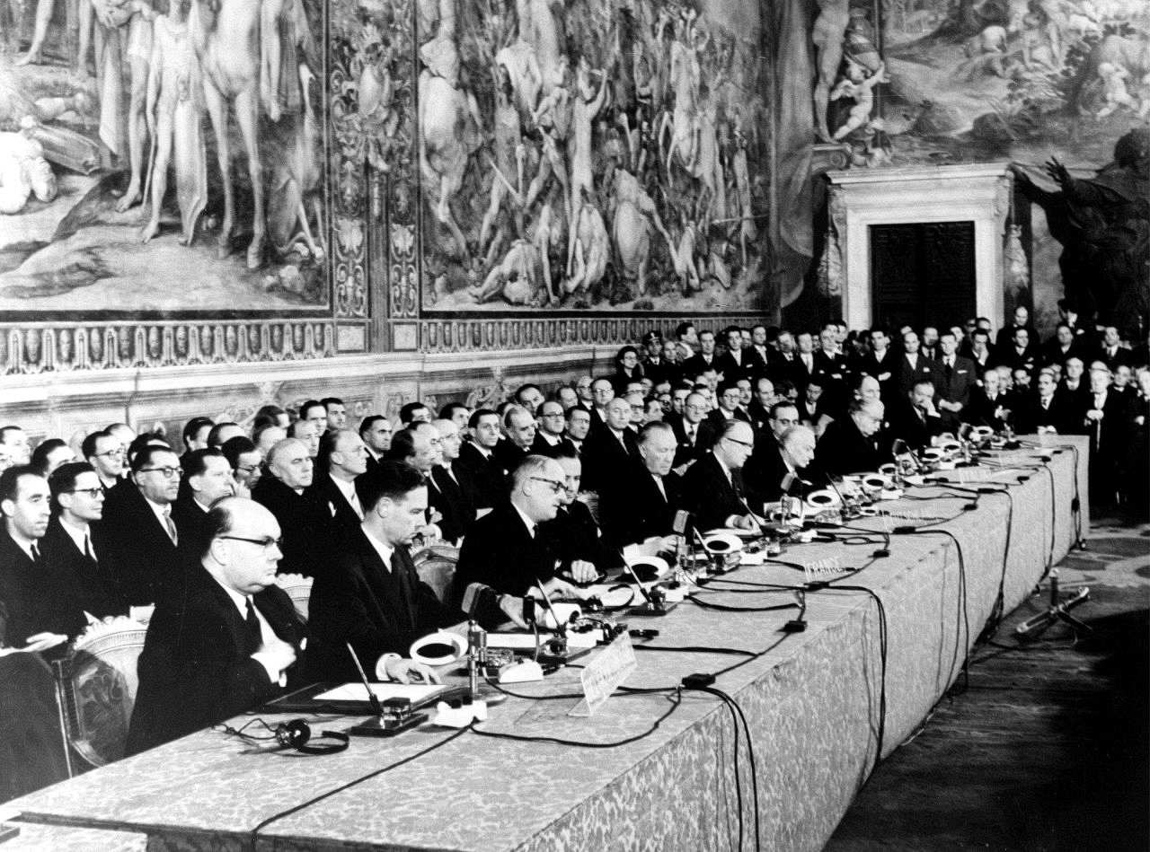 Die unterzeichnenden Staats- und Regierungschefs der Römischen Verträge sitzen an einem langen Tisch. Vor ihnen auf dem Tisch jeweils Mikrofone und Kopfhörer. Hinter ihnen sitzen und stehen weitere Personen. Wände dahinter sind mit Wandmalereien verziert.