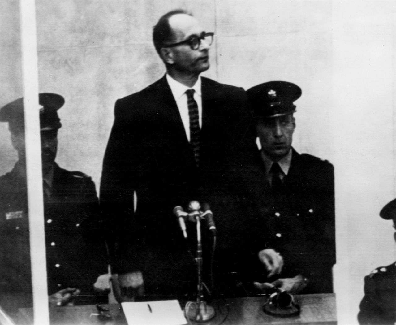 Adolf Eichmann stehend bei seinem Prozess in Jerusalem; vor ihm zwei Mikrofone und hinter ihm sitzend zwei Uniformierte.